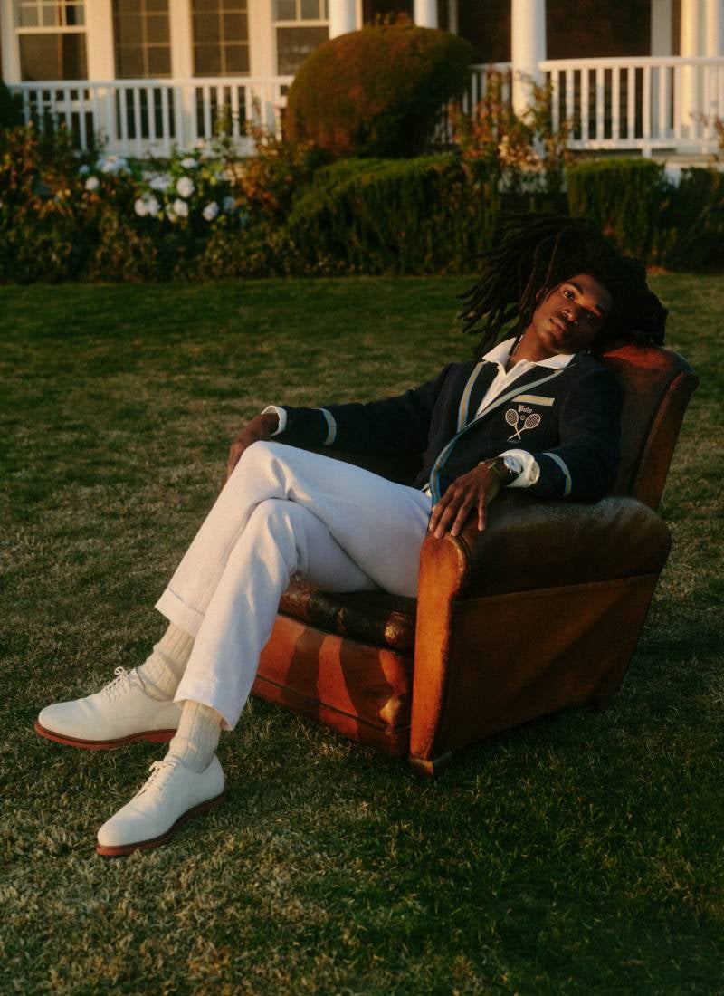 Man in chair on lawn wears navy cardigan and white pants.