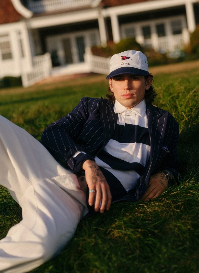 Man lying on lawn wears navy striped blazer and white pants.