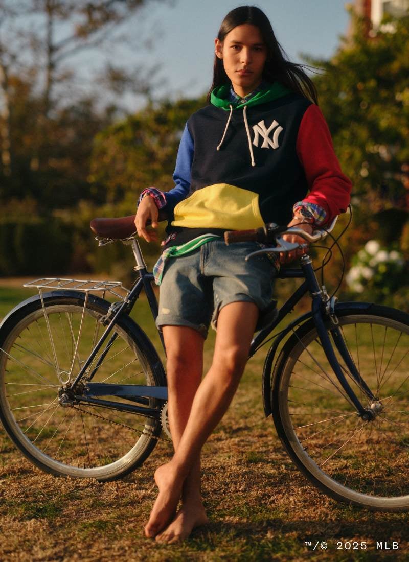 Man leaning against bike wears color-blocked Yankees hooded sweatshirt and jean shorts.
